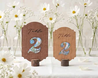 May include: Two scalloped wooden table number signs labeled "Table 2" with iridescent abalone shell inlays. Each sign is mounted on a small, cylindrical wooden base, featuring a dark brown wood finish on the left and a lighter wood finish on the right.