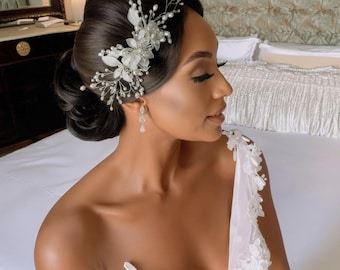 May include: A woman with dark hair styled in an updo, adorned with a delicate floral and pearl hairpiece. She wears a white, off-the-shoulder dress with floral embellishments and dangling earrings. The image is a close-up, focusing on the woman's face and upper body.