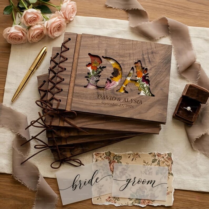 K&ouml;nnte beinhalten: Holz-G&auml;steb&uuml;cher f&uuml;r Hochzeiten mit floral gef&uuml;llten Initialen "DA". Die Namen "David & Alysia" und das Datum "27. September 2026" stehen darunter. Ein goldener Stift, eine Ringbox und Karten mit "bride" und "groom" sind ebenfalls zu sehen.