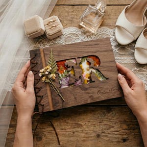 Könnte beinhalten: Ein Holz-Sammelalbum mit den Buchstaben "FR", die ausgeschnitten und mit bunten Trockenblumen gefüllt sind. Der Einband ist mit einer Lederschnur gebunden. Die Worte "Our story 07.15.23" sind auf dem Einband aufgedruckt. Eheringe und Schuhe sind ebenfalls zu sehen.