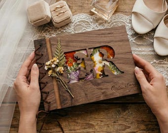 May include: A wooden scrapbook with the letters "FR" cut out and filled with colorful dried flowers. The cover is tied with a leather cord. The words "Our story 07.15.23" are printed on the cover. Wedding rings and shoes are also visible.