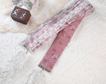 May include: A long, dusty rose ribbon featuring a sheer overlay with a white floral lace trim. The ribbon has a textured, velvet-like appearance and is finished with dangling, clear crystal-like beaded tassels at both ends of the fabric strip.