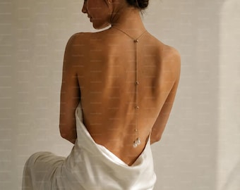 May include: A gold-toned back necklace featuring a delicate chain that drapes down the spine, accented with small metallic beads and finished with two dangling white teardrop-shaped pendants, worn with a sleeveless white satin slip dress.