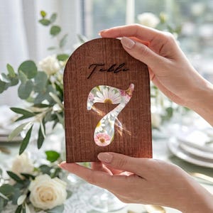 May include: A wooden table number seven with a floral design. The number is cut out, revealing a pattern of white daisies and pink flowers. The word "Table" is written in cursive at the top.