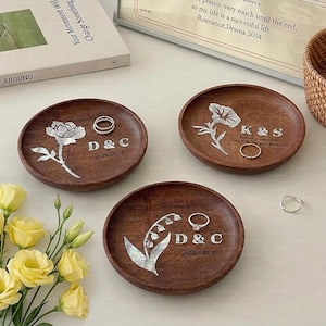 Könnte beinhalten: Drei runde Holz-Ringschalen mit floralen und textlichen Designs. Jede Schale hat ein anderes Blumendesign mit eingelegten Perlmutt-Akzenten. Die Schalen haben Initialen und Daten eingraviert. Ringe sind auf den Schalen platziert.