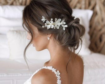 May include: A silver hair comb with delicate white flower and pearl accents. The comb is placed in an updo hairstyle. The person is wearing a white dress with floral shoulder straps. The background includes a bed and woven headboard.