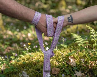 May include: A lavender ribbon featuring a delicate floral pattern, wrapped around two wrists and tied in a knot. The ribbon hangs downward, ending in a clear, faceted crystal pendant that catches the light, creating a decorative accessory.