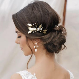 May include: A woman's updo hairstyle featuring a gold and pearl hair accessory. The hairpiece includes white fabric flowers, gold leaves, and small pearls. The woman is also wearing pearl earrings. The image suggests a bridal or formal event.