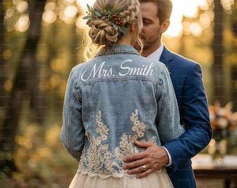 May include: A light blue denim jacket with the name "Mrs. Smith" in white script on the back. The jacket features intricate gold lace detailing and is worn over a cream-colored tulle dress. A man in a blue suit embraces the person wearing the jacket.