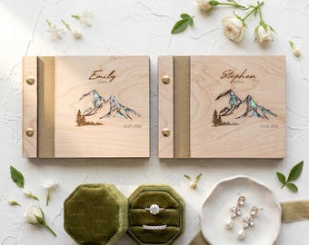 May include: Two square wooden vow books with gold metal binding screws, engraved with the names Emily and Stephen, the word vows, a mountain graphic with iridescent inlay, and the date 10.09.2026. Includes a green velvet ring box and earrings.