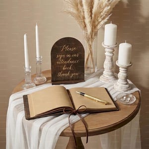 K&ouml;nnte beinhalten: Ein runder Holztisch mit wei&szlig;em Tischtuch zeigt ein G&auml;stebuch, Kerzen und ein Schild mit der Aufschrift "Please sign in our attendance book, thank you". Ein goldener Stift liegt auf dem offenen Buch, und eine kleine Glasschale ist ebenfalls vorhanden.