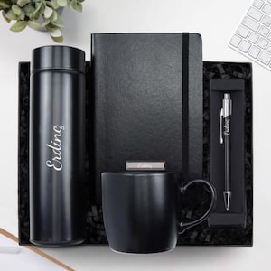 May include: A black gift set featuring a thermos, notebook, mug, and pen, all neatly arranged in a black box. The thermos and pen are inscribed with the name "Erdinç". The set includes a black mug, a notebook with an elastic closure, and a pen.
