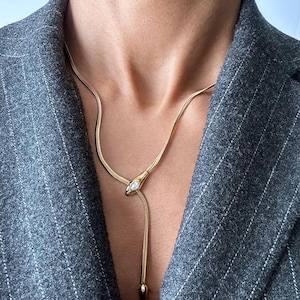 May include: A gold snake chain necklace with a clear stone accent. The chain is adjustable and has a small, pointed pendant at the end.