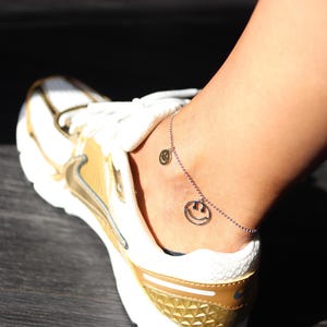 Könnte beinhalten: Ein silbernes Fußkettchen mit zwei Smiley-Anhängern wird an einem Knöchel getragen. Der Knöchel befindet sich neben einem weißen und goldenen Sportschuh. Der Schuh hat weiße Schnürsenkel und eine goldene Ferse.