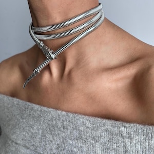 Op de afbeelding: Een zilverkleurige choker-ketting met een ontwerp met drie strengen. De ketting wikkelt zich om de nek en heeft een decoratieve, puntige hanger. De sieraden zijn gemaakt van een glanzend, metalen materiaal.