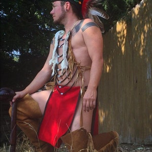 May include: A person wearing a red and brown traditional Native American outfit, including a feathered headdress, beaded necklace, and leather skirt. They are holding a wooden staff and are kneeling in front of a wooden fence.