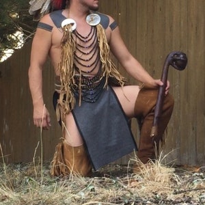 May include: A man wearing a black and blue beaded headdress, a grey skirt, and brown leather boots. He is holding a wooden staff with a carved head. He is standing in front of a wooden fence.