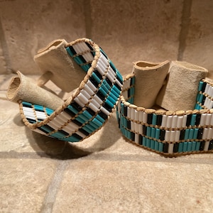 May include: Two beaded bracelets with a geometric pattern of turquoise, black, and white beads. The bracelets are made of leather and have a suede backing.