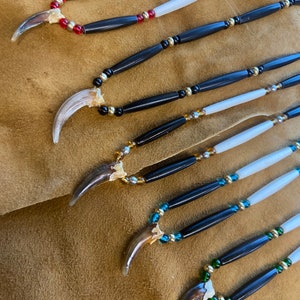 May include: Five necklaces with bear claws as pendants. Each necklace features black and white beads, with different colored accent beads. The necklaces are arranged in a row on a brown suede surface.