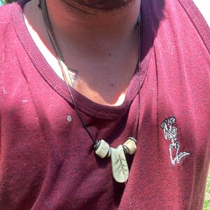 May include: A close-up of a person wearing a maroon tank top with a necklace featuring a carved leaf pendant and two round beads. The pendant has a small tree design carved into it. The tank top has a small embroidered skeleton design on the right side.