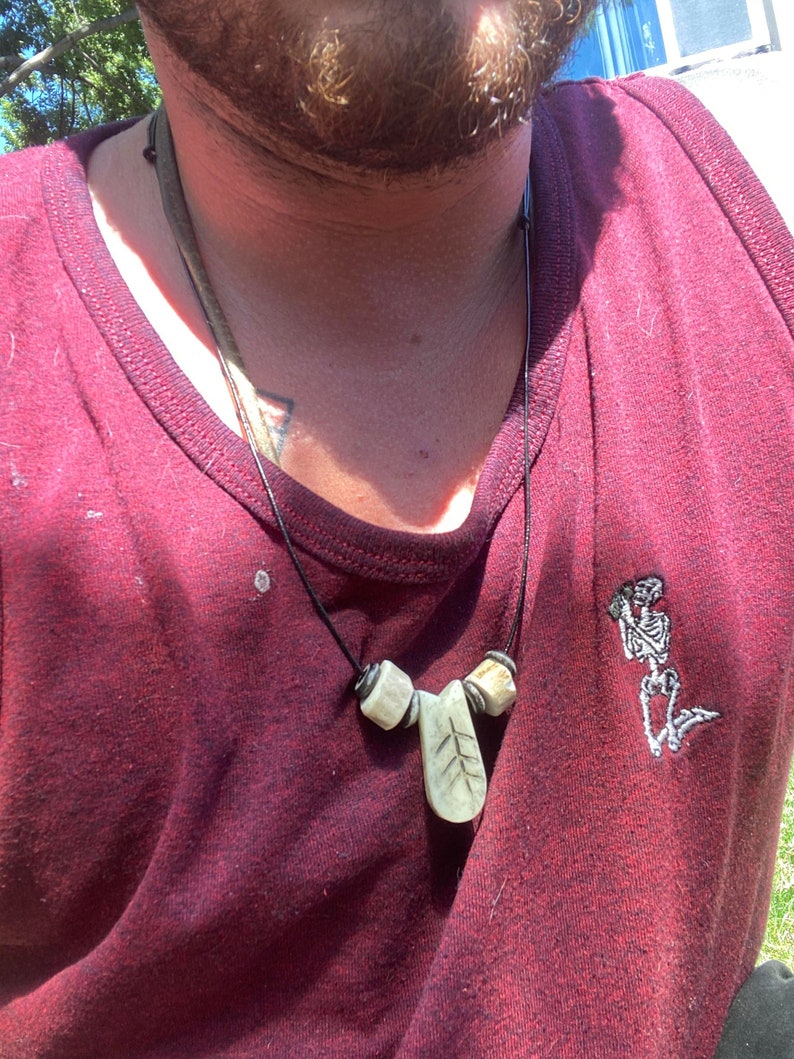 May include: A close-up of a person wearing a maroon tank top with a necklace featuring a carved leaf pendant and two round beads. The pendant has a small tree design carved into it. The tank top has a small embroidered skeleton design on the right side.