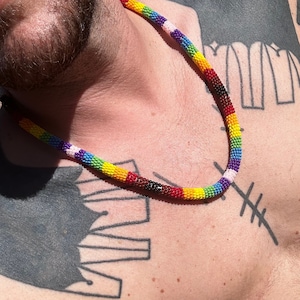 May include: A close-up of a person wearing a rainbow beaded necklace. The necklace is made of small, colorful beads and is worn around the neck. The person has a tattoo on their chest.