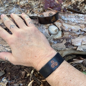 May include: Two leather bracelets with a Celtic knot design. The bracelet on the left is brown and the bracelet on the right is black.