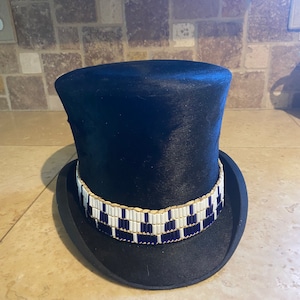 May include: A black top hat with a white and blue beaded band. The hat is made of a smooth, black fabric.