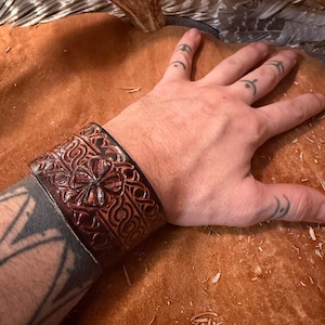 May include: A wide, dark brown leather cuff bracelet with an embossed floral and scroll design. The cuff is worn on a wrist and features intricate details. The leather has a rich, textured appearance, suggesting a handcrafted item. The bracelet is a statement piece.