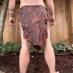 May include: A brown leather skirt with ties on the sides. The skirt is worn by a person with tattoos on their arms and back.