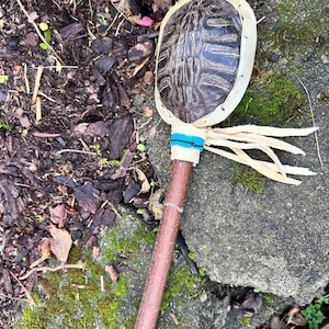 May include: A handmade turtle shell rattle with a wooden handle. The rattle features a dark brown turtle shell, a white leather rim, and a handle wrapped with blue and white thread. The handle is made of a brown stick with a natural finish.