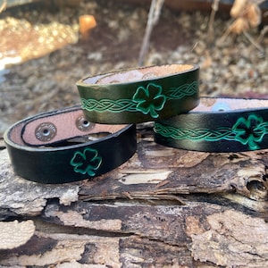 May include: Two black leather bracelets with green shamrock designs and Celtic knot patterns. The bracelets have snap closures.