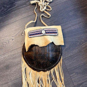 May include: A brown leather bag with a beaded strap and fringe. The bag is made from a turtle shell and has a beaded design.