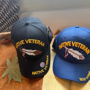 May include: Two baseball caps, one black and one navy blue, are displayed. Both caps feature the text "NATIVE VETERAN" in gold embroidery, along with a feather design. The black cap has a brown and white feather, while the blue cap has a white and red feather.