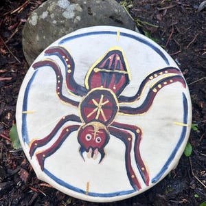 May include: A round, off-white drum with a painted spider design. The spider has a red body and legs, with gold accents. A blue line borders the edge of the drum.