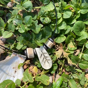 May include: A white bone pendant with a leaf design hangs from a black cord with white bone beads. The pendant is lying on a bed of green leaves.