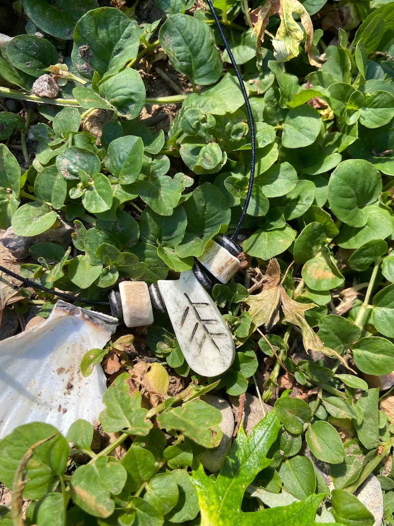 May include: A white bone pendant with a leaf design hangs from a black cord with white bone beads. The pendant is lying on a bed of green leaves.