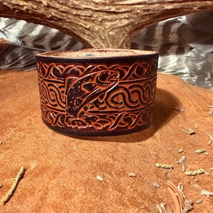 May include: A brown leather cuff bracelet with a detailed embossed design. The design features a leaping fish and Celtic knot patterns. The bracelet is approximately 2 inches wide.