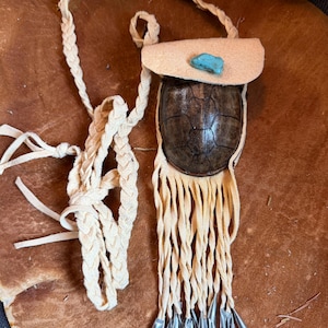 May include: A small, handcrafted pouch with a turtle shell base and a cream-colored leather flap. The pouch features a turquoise stone accent and cream-colored fringe with silver-toned metal tips. A braided cream-colored leather strap is attached.