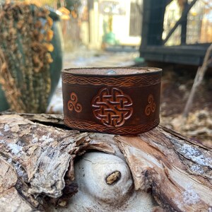 May include: A brown leather bracelet with a Celtic knot design and a triple spiral pattern. The bracelet is on a wooden surface.