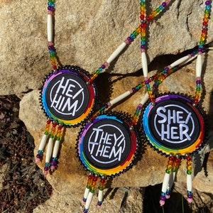 May include: Three beaded necklaces with circular pendants featuring the words "He/Him", "She/Her", and "They/Them" in white text on a black background. The pendants are surrounded by rainbow-colored beads.
