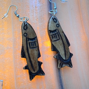 May include: A pair of wooden fish earrings with silver-colored hooks. The earrings are a light brown color with dark brown etched details, including eyes and a design on the body. The fish have a star-shaped tail.