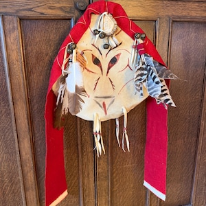 May include: A Native American-style mask with a red felt hood and streamers. The mask is painted with a face design and adorned with feathers, beads, and a small antler piece. The mask is hanging on a wooden door.