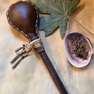 May include: A handcrafted, brown leather rattle with a dark wooden handle. The rattle head is stitched with white thread. Silver-colored metal tassels hang from the handle. A large green leaf and a seashell with dried herbs are also in the image.