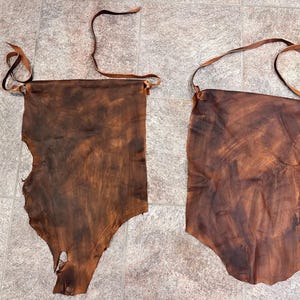 May include: Two pieces of brown leather with a rough texture and a natural, rustic look. The leather is cut into a rectangular shape with a rounded bottom and has a hole at the top for hanging.