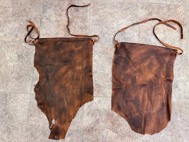 May include: Two pieces of brown leather with a rough texture and a natural, rustic look. The leather is cut into a rectangular shape with a rounded bottom and has a hole at the top for hanging.