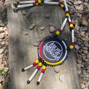 May include: A beaded necklace with a round black patch that says "Ask Me About My Pronouns". The patch is surrounded by red, yellow, and black beads. The necklace is made with white bone beads and black beads.