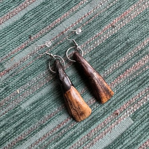 May include: A pair of wooden earrings with a black and brown wood grain pattern. The earrings are teardrop shaped and have silver hooks.