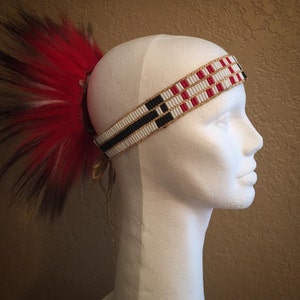 May include: A beaded headband with red, white, and black beads. The headband is worn on a white mannequin head. A red, black, and white feather headdress is attached to the side of the headband.