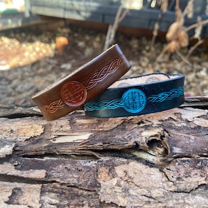 May include: Two leather bracelets with Celtic knot designs. One is brown, the other is black with a turquoise design. The bracelets are on a piece of weathered wood. The bracelets are a unique accessory.